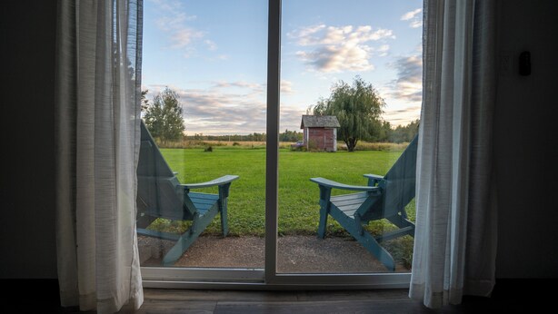 A room with a large sliding glass door leading to an outdoor area with two blue Adirondack chairs and a small red shed.