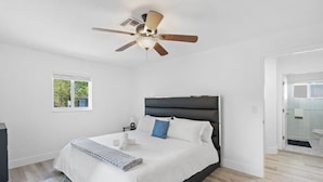Ein modernes Schlafzimmer mit einem großen Bett, einem Deckenventilator, einem Fenster und einem durch eine offene Tür einsehbaren Badezimmer.