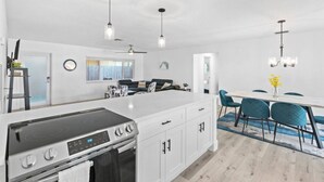 Eine moderne Küche mit Kochinsel, weißen Schränken und integriertem Backofen. Es gibt einen Essbereich mit Tisch und Stühlen sowie einen Deckenventilator.