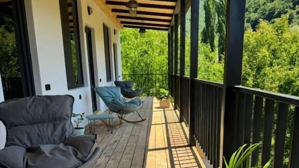 A wooden deck with outdoor furniture, a potted plant, and a view of lush greenery. - Debatoun (Debet)