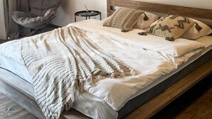A bedroom with a wooden bed frame, a large bedspread, a chair, a small table, and a hanging light fixture. - Debatoun (Debet)