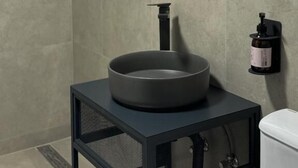 A modern bathroom with a round sink on a black pedestal, a wall-mounted mirror, and a small shelf with toiletries. - Debatoun (Debet)