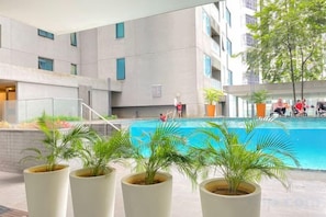 A swimming pool area with potted plants, a building with multiple windows, and people in the background. - Summer Suites KLCC by Luna (Kuala Lumpur)