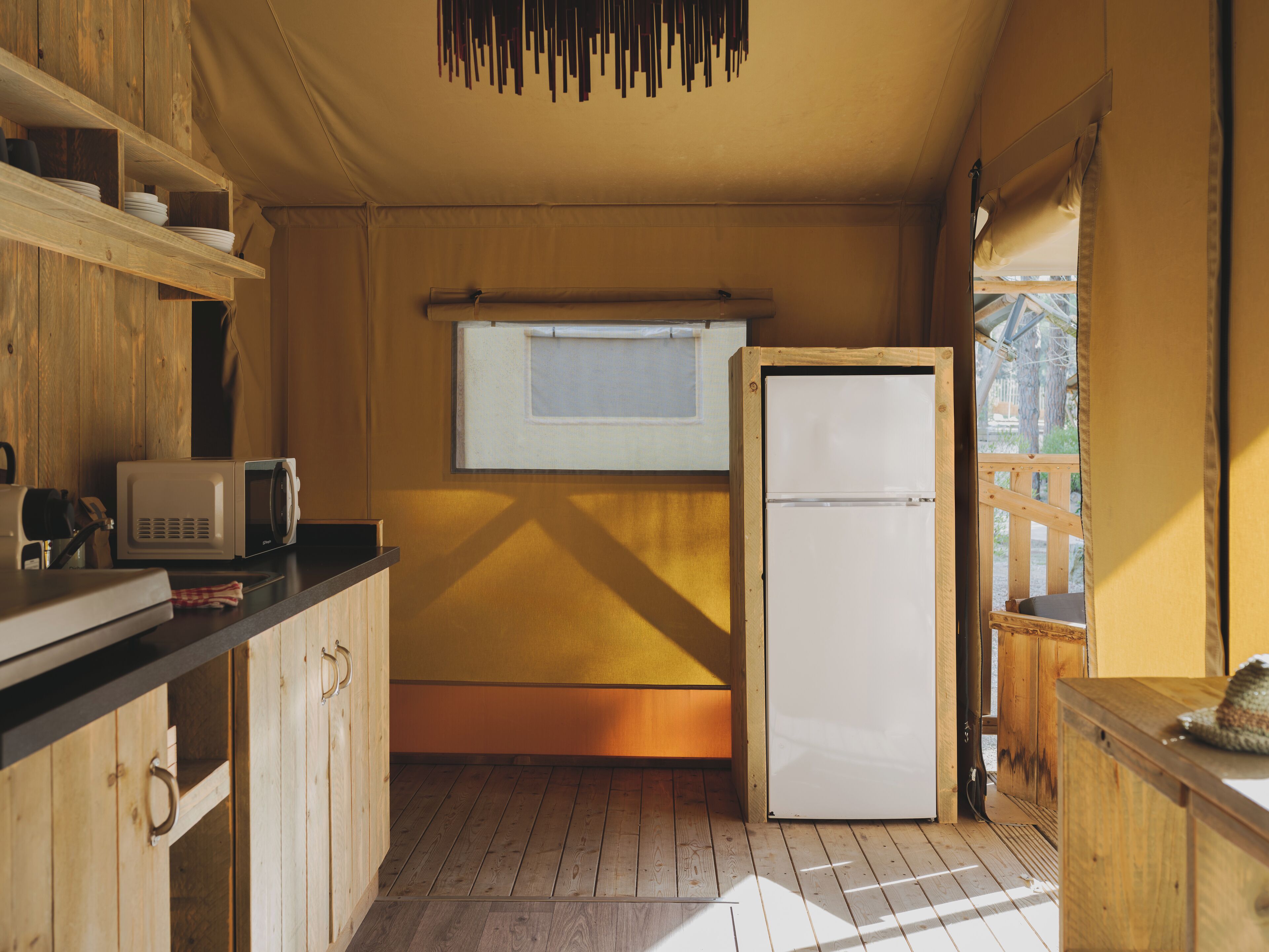 A compact kitchen with wooden cabinets, a microwave, a refrigerator, and a window with a view of the outdoors.