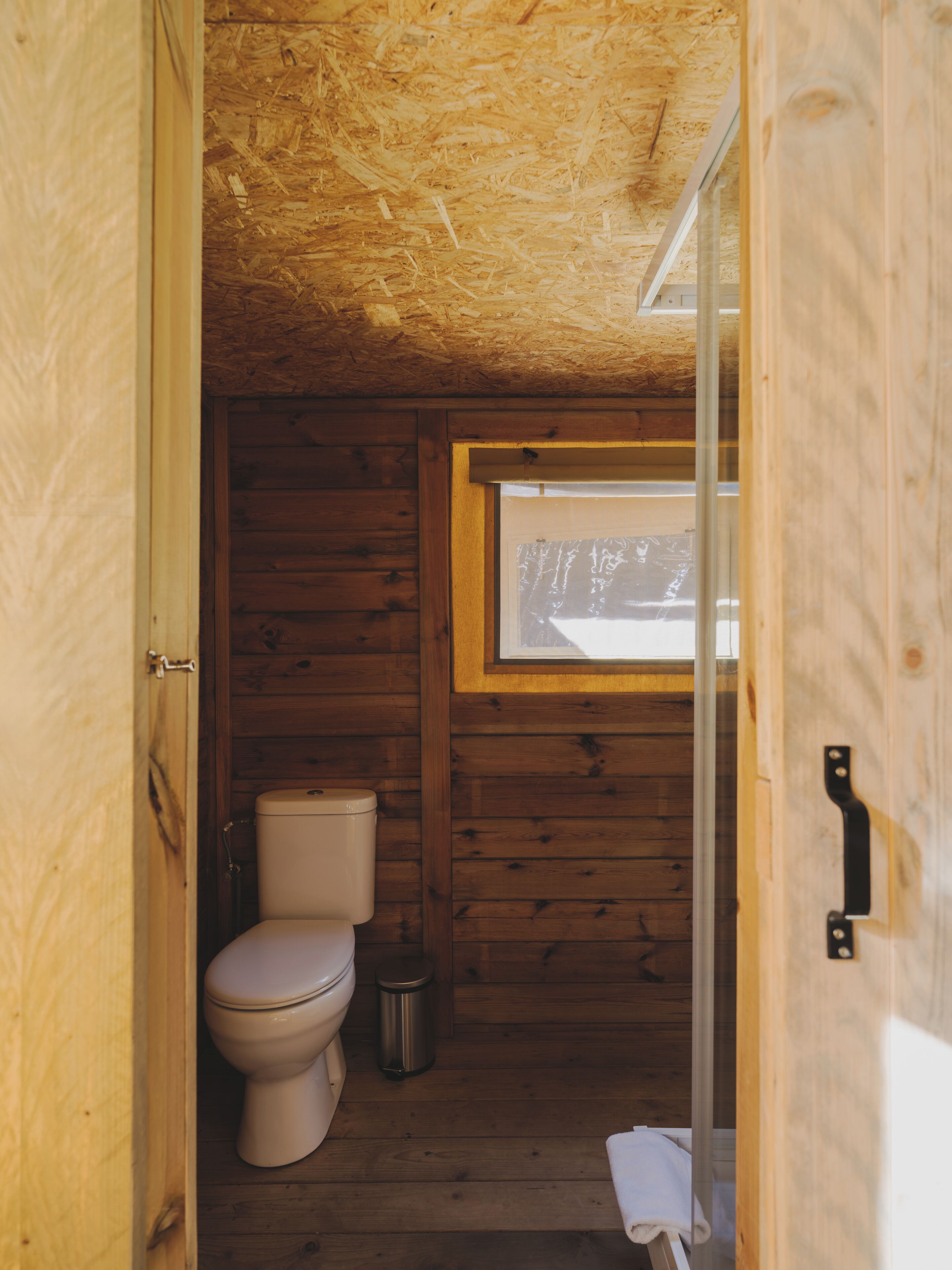 Une petite salle de bain de style cabane en bois, équipée de toilettes, d’une poubelle et d’une fenêtre laissant entrer la lumière naturelle.