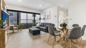 Un salon moderne avec un canapé d’angle gris, une table à manger en verre entourée de chaises grises et un bureau en bois agrémenté d’une plante.