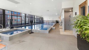 Une piscine intérieure moderne avec des parois en verre transparent, un sol carrelé et un espace douche agrémenté d’une plante en pot.