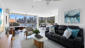A modern living room with a black leather sofa, a wooden coffee table, and a view of the city skyline through large windows. - Stylish 2-bed in Surfers Paradise With River Views (Gold Coast)