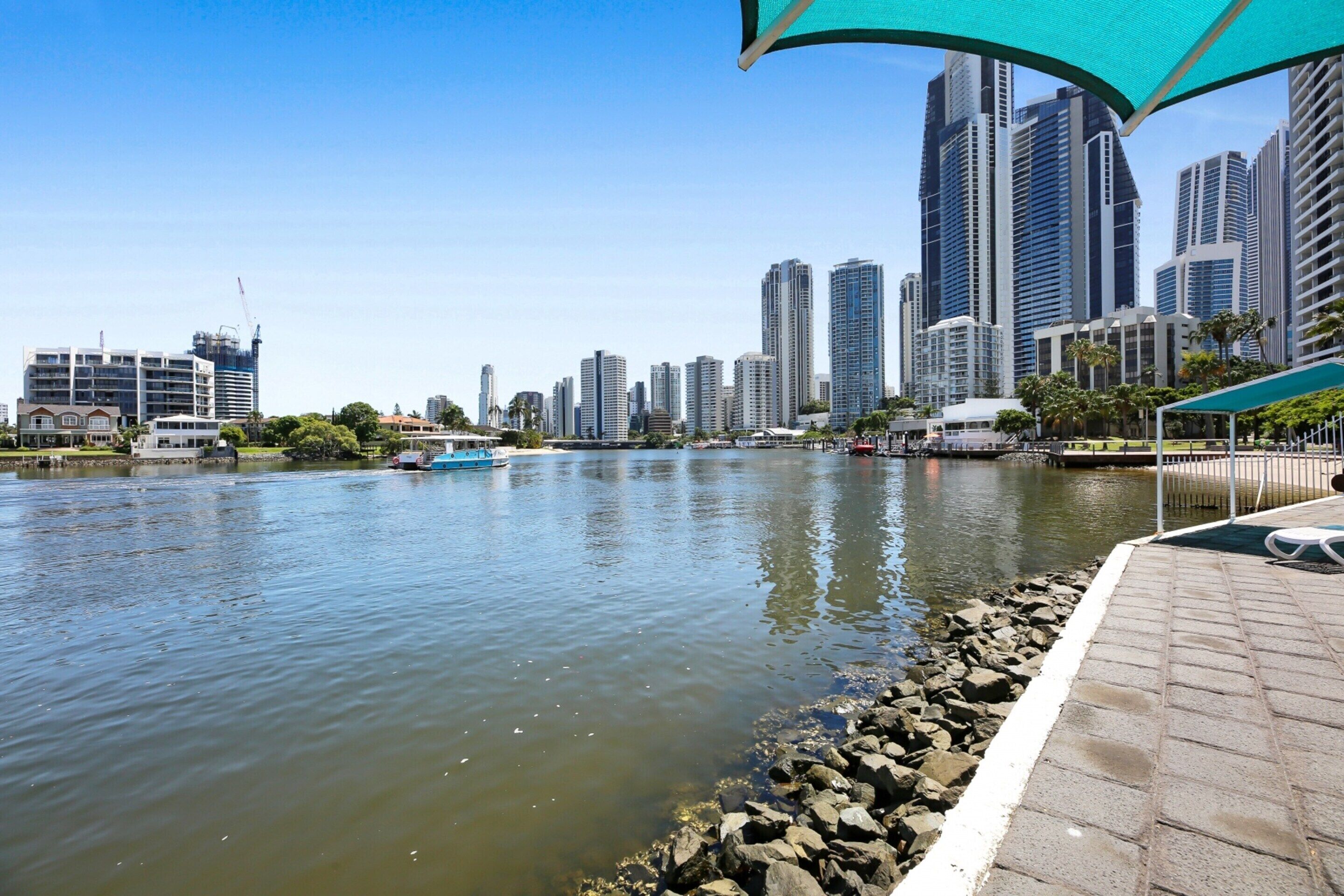 A river with a city skyline in the background, a boat on the water, and a paved walkway with a canopy.