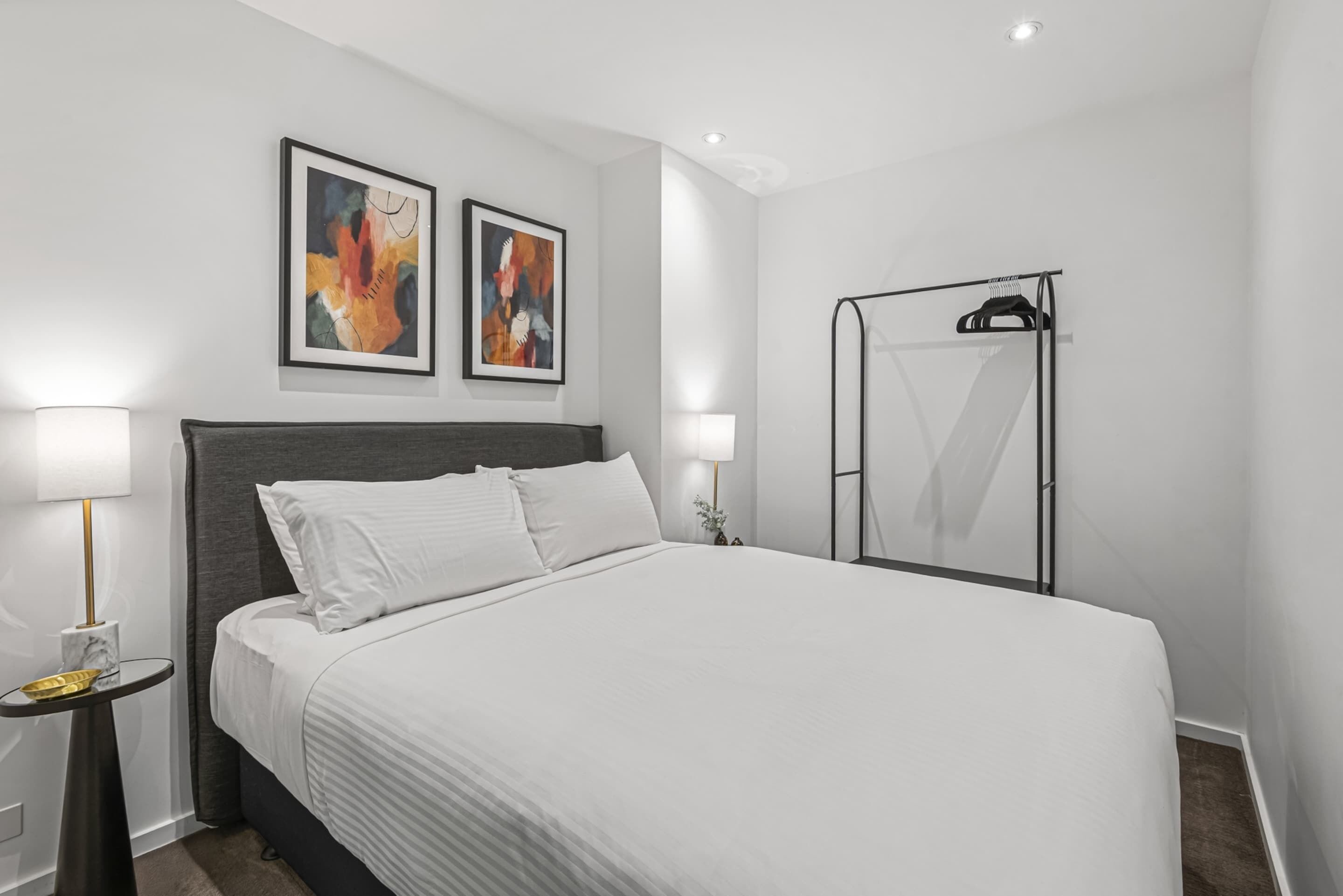 A modern hotel room with a large bed, two bedside lamps, a wardrobe, and abstract artwork on the wall.