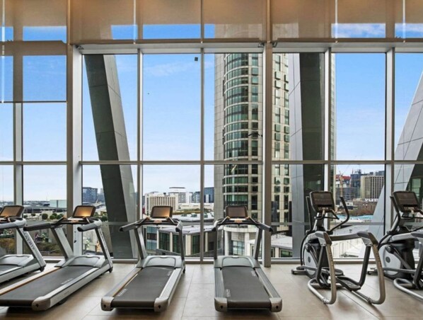 Et fitnesscenter med løbebånd og crosstrainere, store vinduer og udsigt til en høj bygning.
