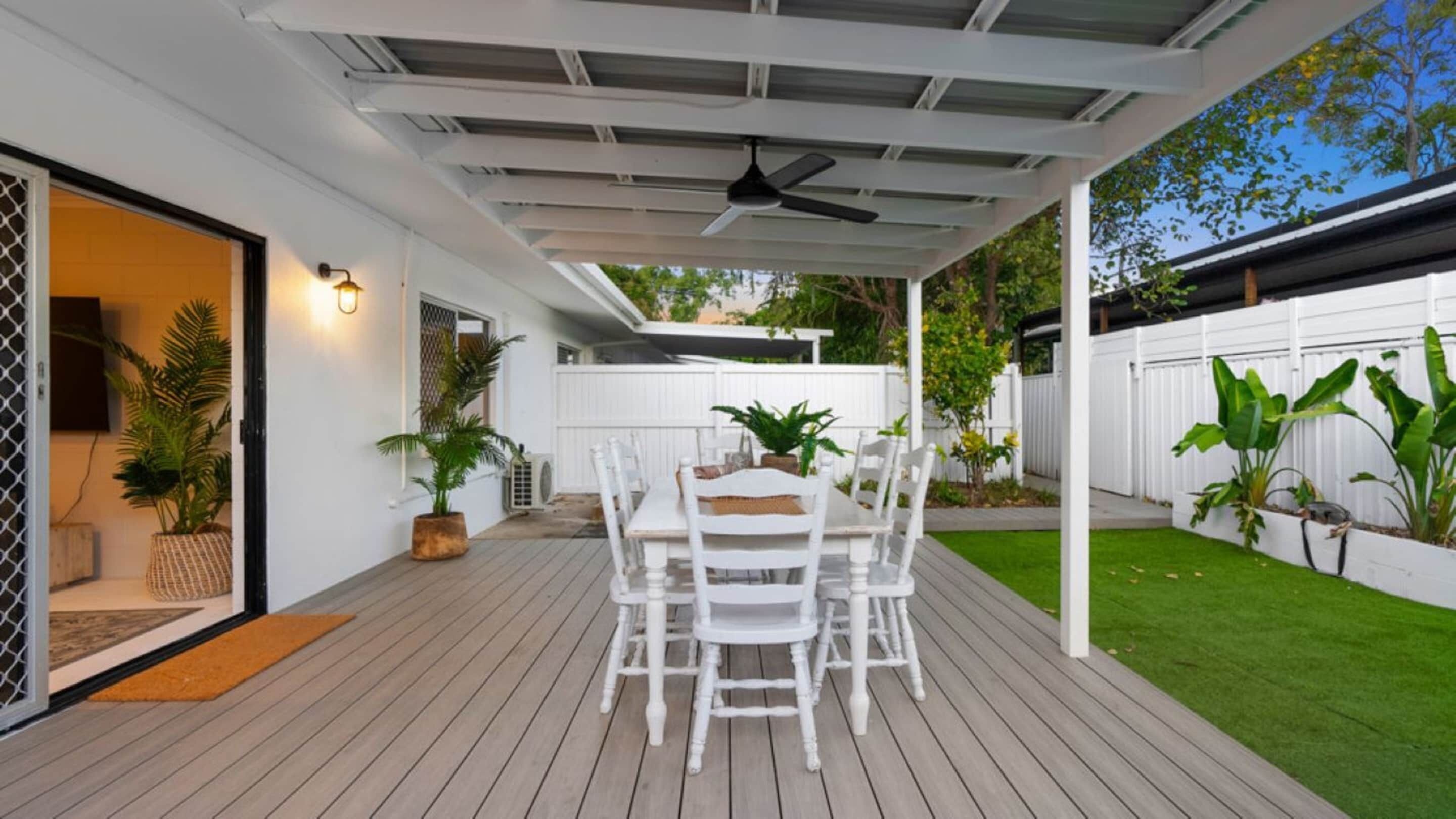 A covered patio with a wooden deck, white outdoor furniture, and potted plants.