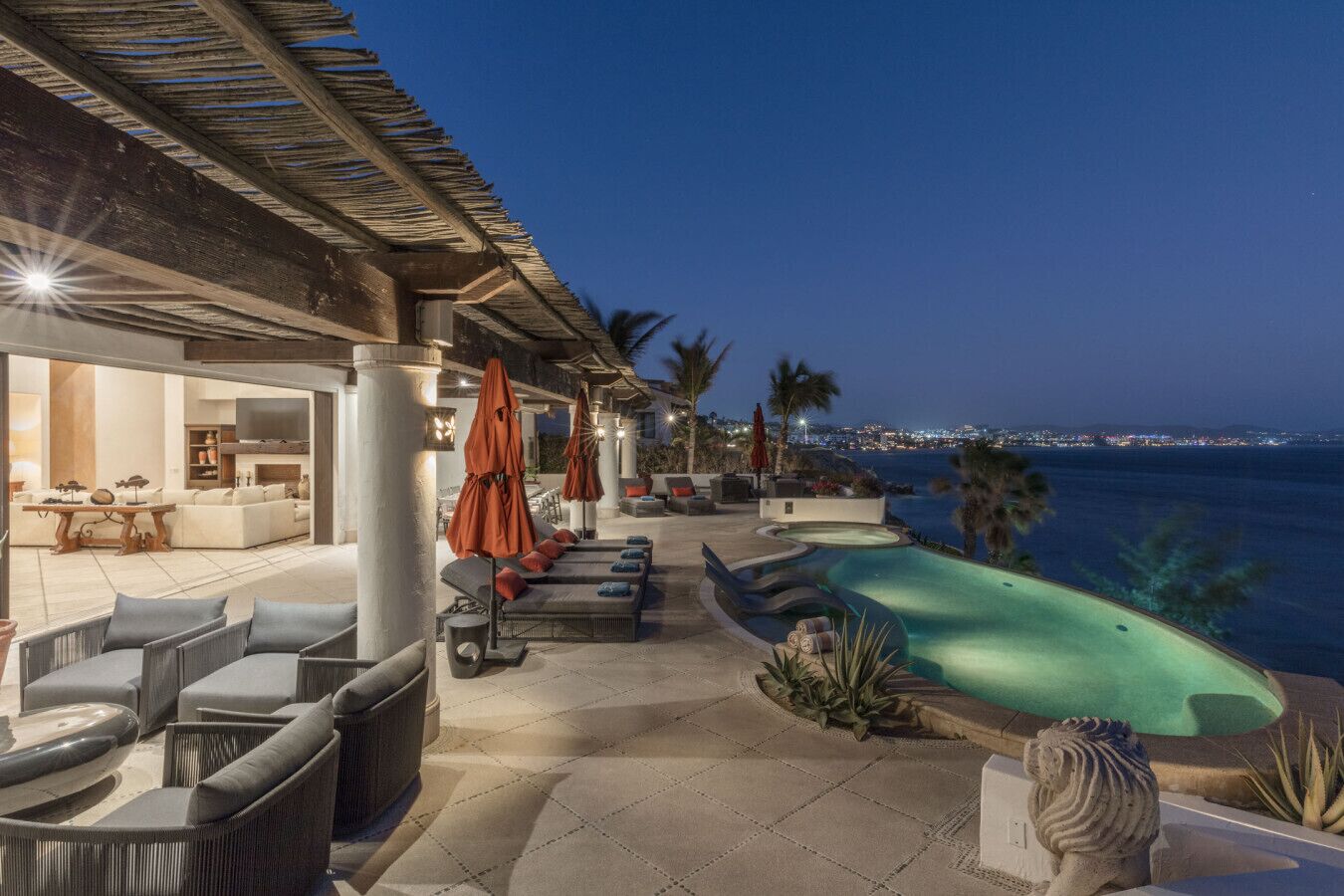 A poolside area with lounge chairs and umbrellas, a view of the ocean, and a building with a covered patio.