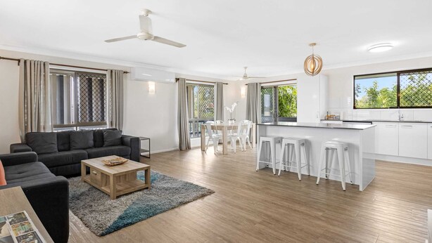 A modern living room with a sofa, coffee table, and a dining area with chairs and a table. - Spacious 3-bed Home With Large Balcony (Hervey Bay)