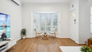 Ein modernes Wohnzimmer mit Holzboden, einem runden Tisch mit Stühlen, einem Fernseher auf einem weißen Schrank und einem großen Fenster mit Rollläden.