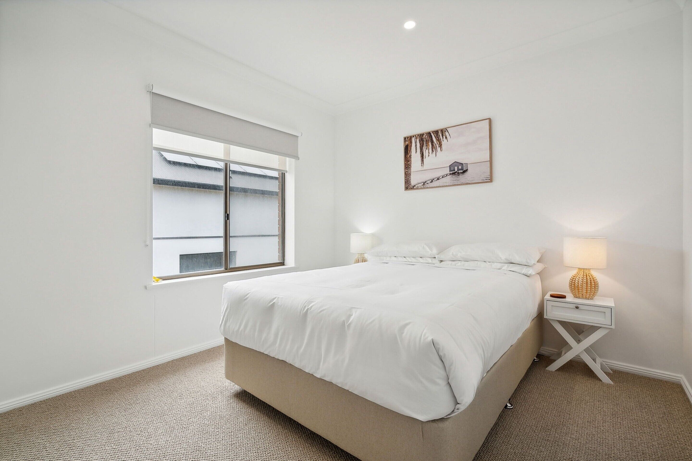 Une chambre à coucher comprenant un lit, une table de chevet, une lampe, une fenêtre avec des stores et un tableau accroché au mur.