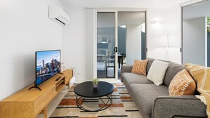 A modern living room with a flat-screen TV, a round coffee table, a grey sofa with patterned cushions, and a wooden side table. - Comfy 2-bed With Pool and Secure Parking (Brisbane)
