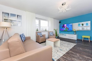 Ein modernes Wohnzimmer mit einem beigefarbenen Sofa, zwei Sesseln, einem Couchtisch, einem Fernseher und einer blauen Akzentwand.