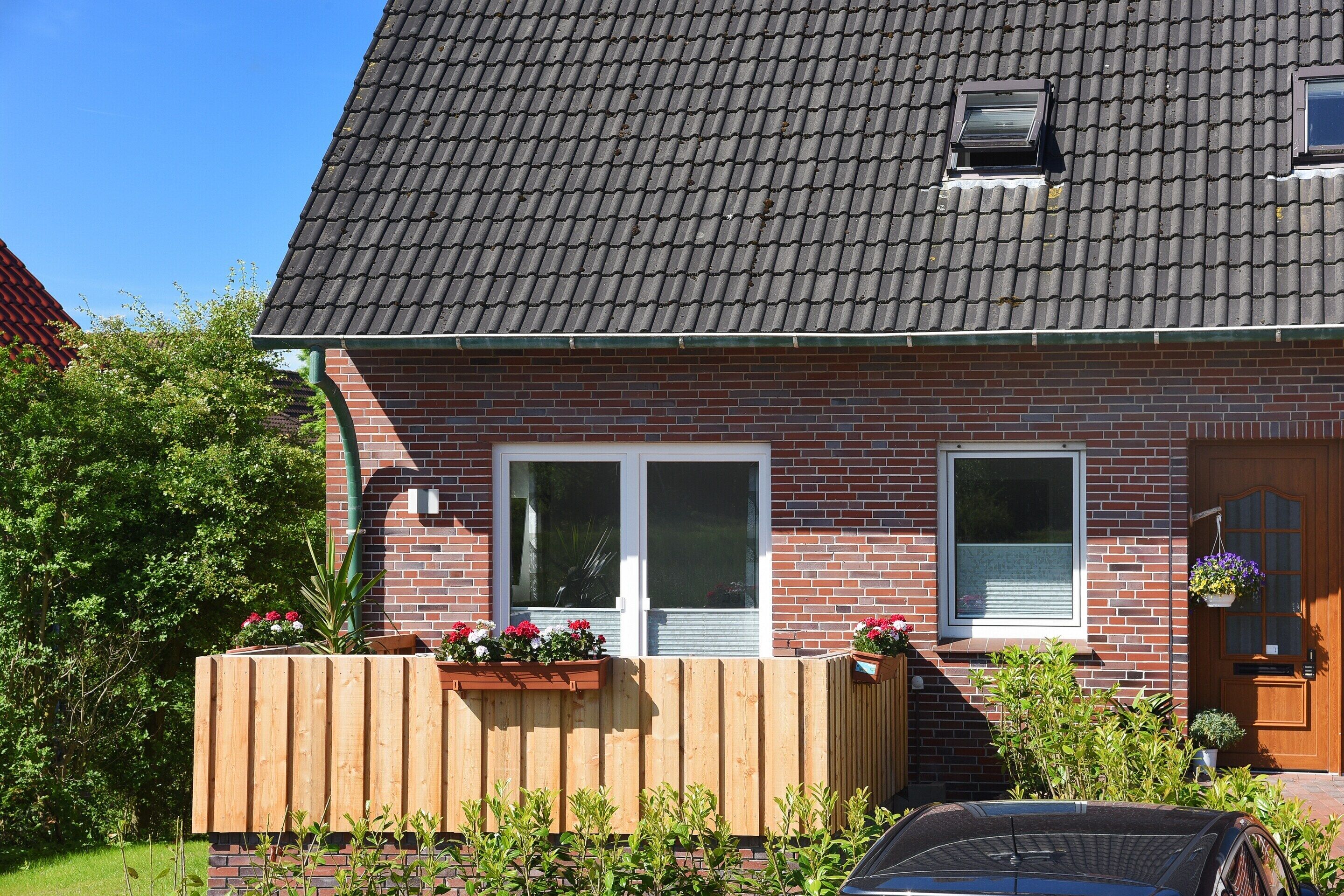 A brick house with a tiled roof, two windows, a wooden fence, and a car parked in front.