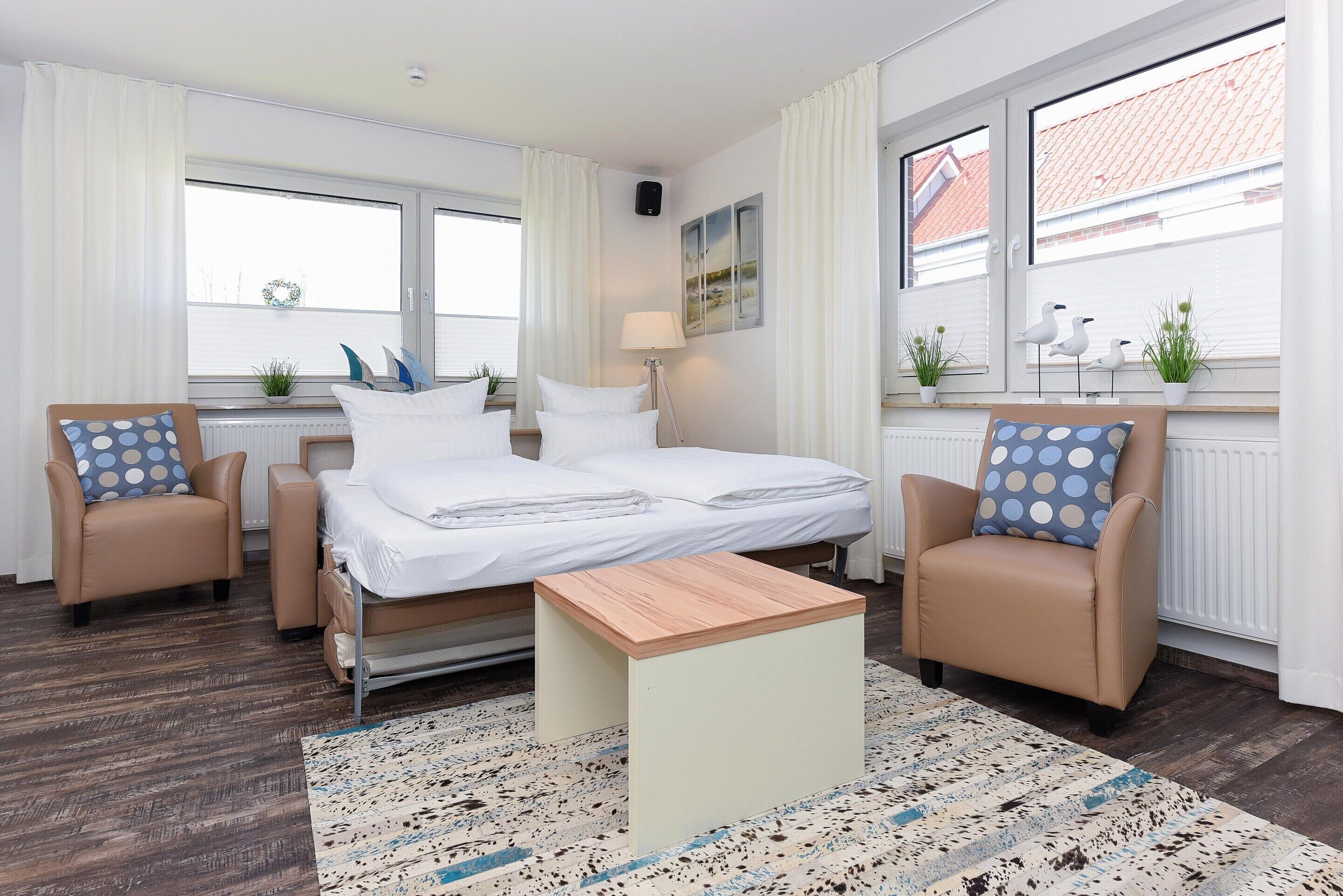 Ein Hotelzimmer mit einem Bett, zwei Sesseln, einem kleinen Tisch und großen Fenstern mit Vorhängen.