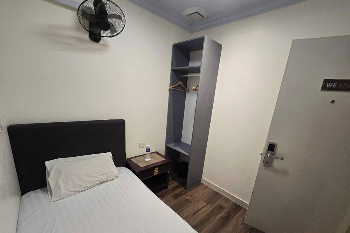 Ein Hotelzimmer mit Bett, Nachttisch, Ventilator, Kleiderschrank und einer Tür mit einem Schild.