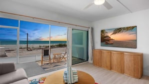 Una habitación con un amplio ventanal corredizo que da a una vista de la playa, un armario de madera, un cuadro con escena costera y una pequeña zona de estar con una mesa.