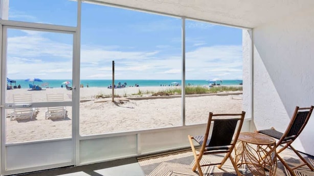 Vista a la playa desde una habitación con ventanas amplias, muebles de mimbre y un tapete estampado.