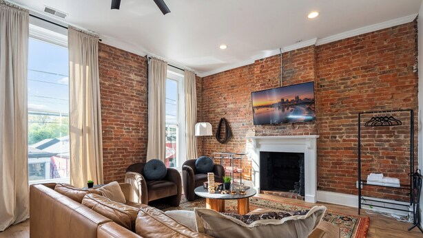 A living room with a brick wall, leather sofa, and a fireplace. - Stable #1 ~Walkable, BBQ, Fire Pits, Mini Golf (Louisville)