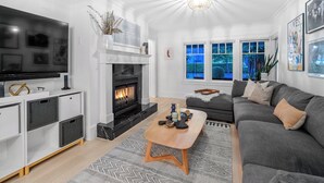 A modern living room with a fireplace, a grey sofa, a wooden coffee table, and a TV. - Charming 4-Bedroom Home in Peaceful Parkside Setting with Hot Tub & Sauna (North Vancouver)