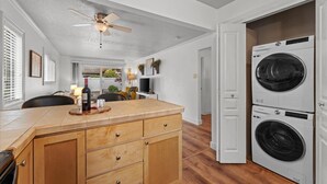 A laundry room with stacked washers and dryers, a wooden cabinet with drawers, a ceiling fan, and a view into the adjacent living area. - Ground Level Condo by Sunset High School (Portland)