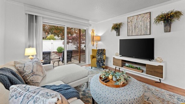 A living room with a sofa, ottoman, and a TV on a white stand. There are plants on the wall and a window with a view of trees. - Ground Level Condo by Sunset High School (Portland)