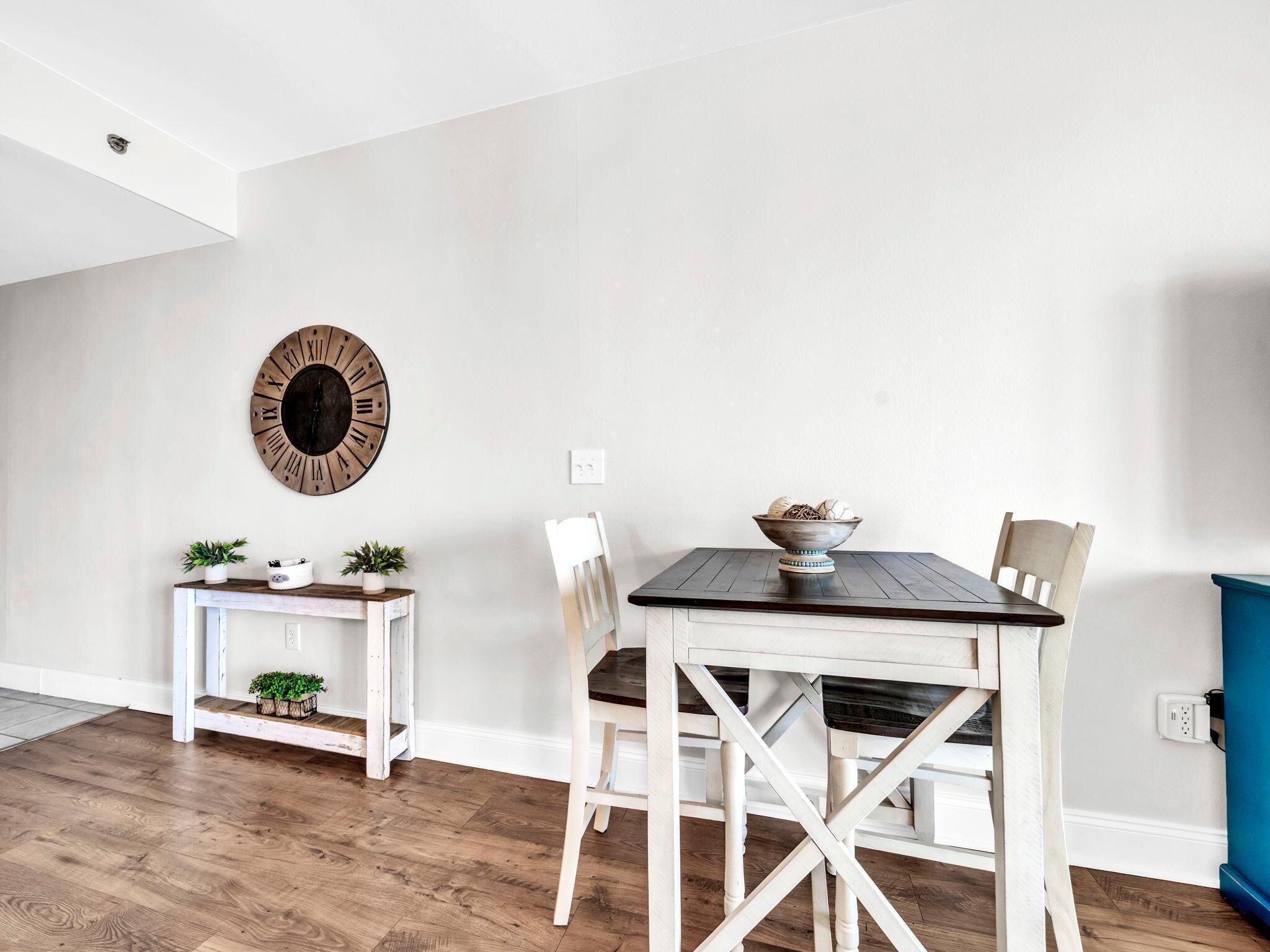 Un coin repas avec une table et des chaises en bois, une horloge murale et une petite table d’appoint agrémentée de plantes.