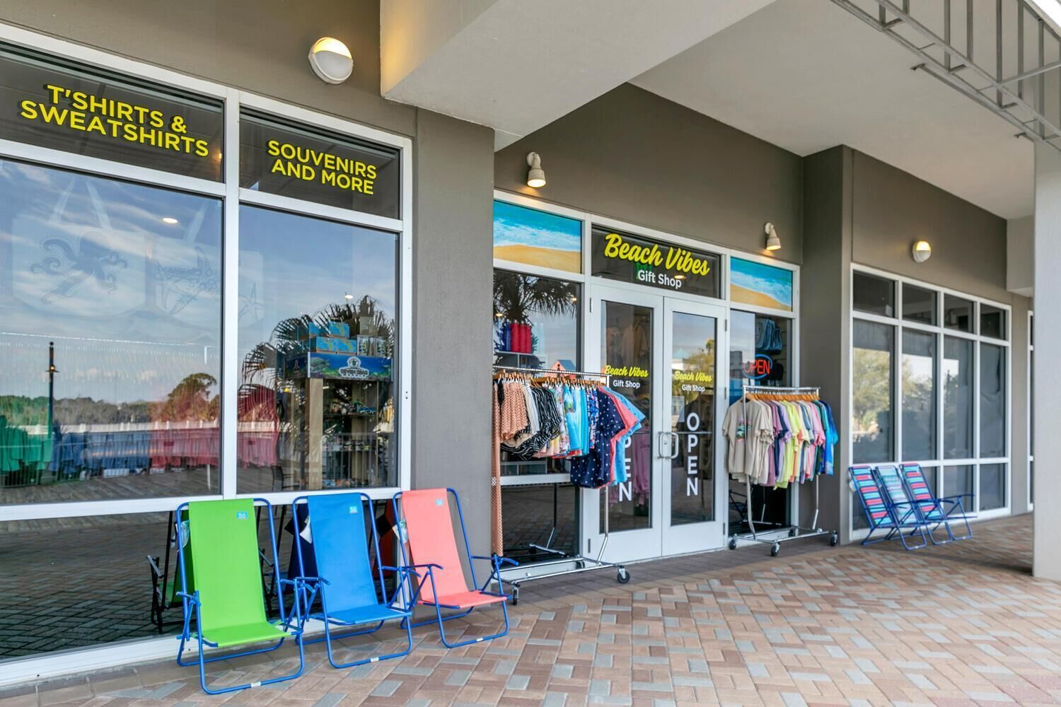 A storefront with 'T'SHIRTS & SWEATSHIRTS' and 'SOUVENIRS AND MORE' signs, a 'Beach Vibes Gift Shop' sign, and colorful chairs outside.