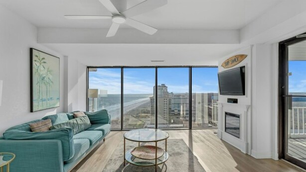 A modern living room with a teal sofa, a glass coffee table, and a city view through large windows. - Forest Dunes 1802- Ocean View 3BR/2.5BA- Pool (Myrtle Beach)