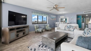 A modern living room with a flat-screen TV, a ceiling fan, and a large window with a view. - Regency Towers 907- Oceanfront 3BR- Pool (Myrtle Beach)