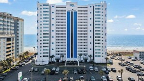 A high-rise hotel with a central blue stripe, parking lot, and a beach view. - Regency Towers 907- Oceanfront 3BR- Pool (Myrtle Beach)
