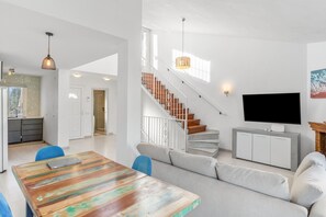 A modern living room with a staircase, a flat-screen TV, and a dining area with a wooden table.