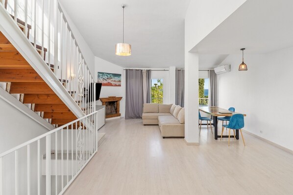 A spacious living area with a wooden staircase, a sofa, a dining table with chairs, and a view of the outdoors.