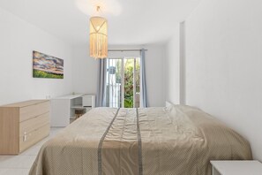 A bedroom with a bed, a desk, a dresser, a hanging light fixture, and a painting on the wall.