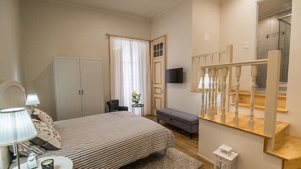 Una camera da letto con un letto, lampade da comodino, una sedia, un tavolino con lampada, una TV, una finestra con tende, una panca e una scala con corrimano.