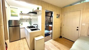 A compact kitchen with a refrigerator, stove, sink, and a small trash bin. There is a door leading to a bathroom and a bed in the room. - Charming cabin in Pāhoa for a peaceful getaway (Pāhoa)