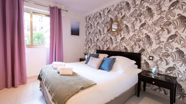A bedroom with a large bed, a nightstand, a window with purple curtains, and a wall with a bird and plant wallpaper. - Charming Riviera 2F - One Bedroom Apartment, Sleeps 4 (Antibes)