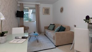 A living room with a beige sofa, a glass coffee table, a white armchair, a TV mounted on the wall, and a window with curtains. - Charming Riviera 2F - One Bedroom Apartment, Sleeps 4 (Antibes)