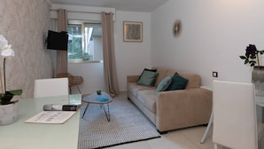 Een woonkamer met een beige bank, een glazen salontafel, een witte fauteuil, een aan de muur bevestigde televisie en een raam met gordijnen.