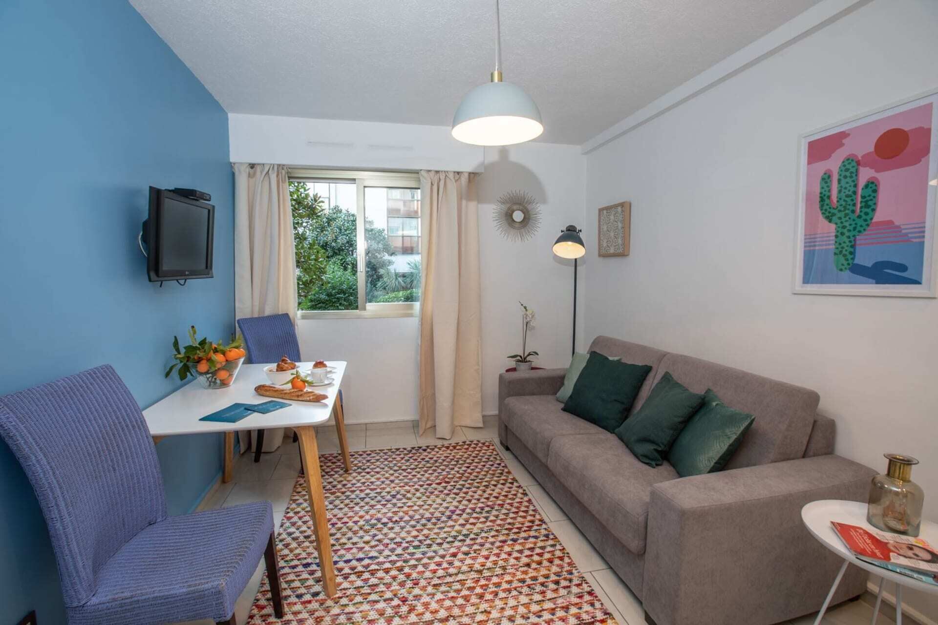 A compact living space with a dining table, a sofa, a chair, a patterned rug, and a window with curtains.