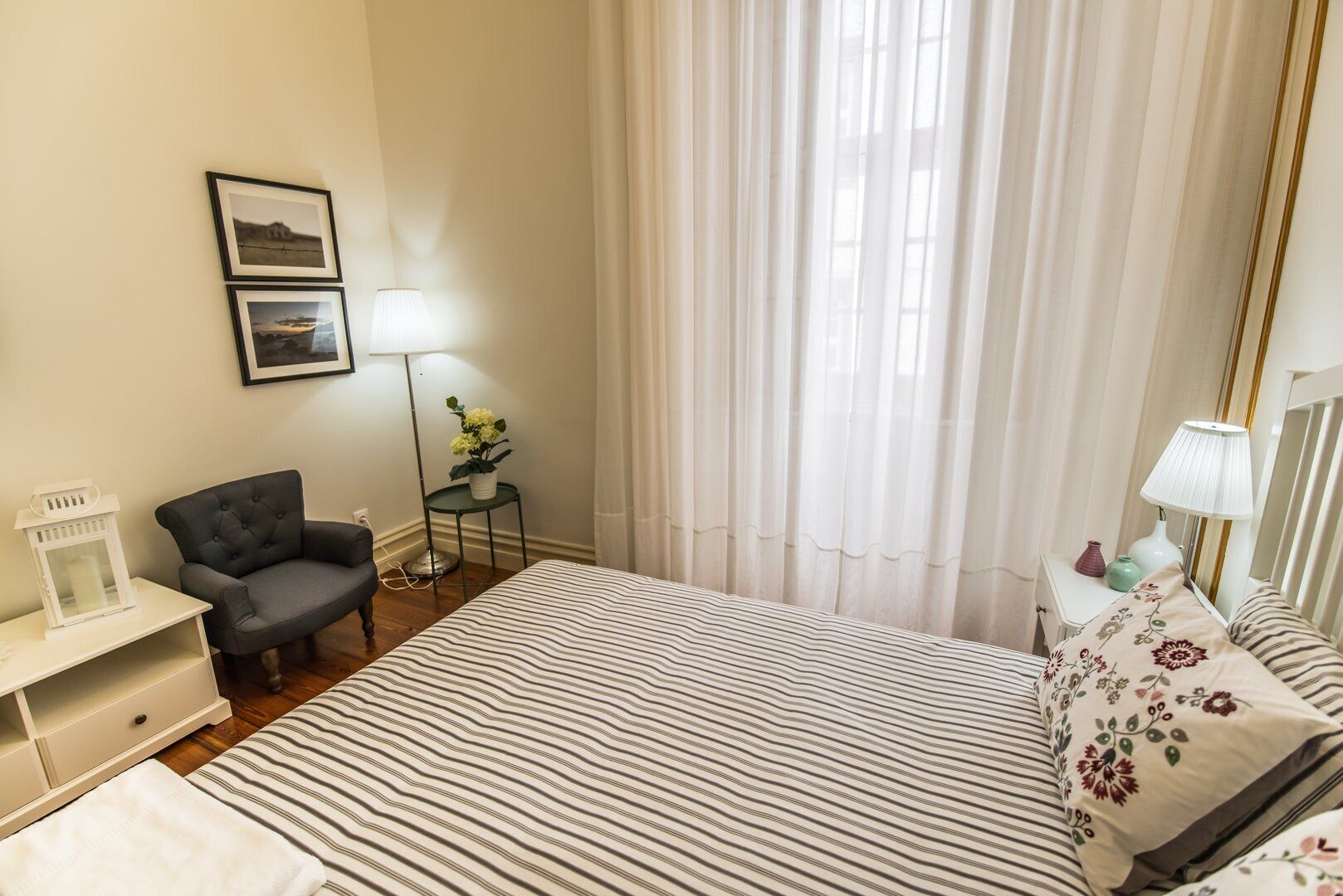 Une chambre d’hôtel avec un lit, une chaise, une table de chevet, une lampe et une fenêtre avec des rideaux.