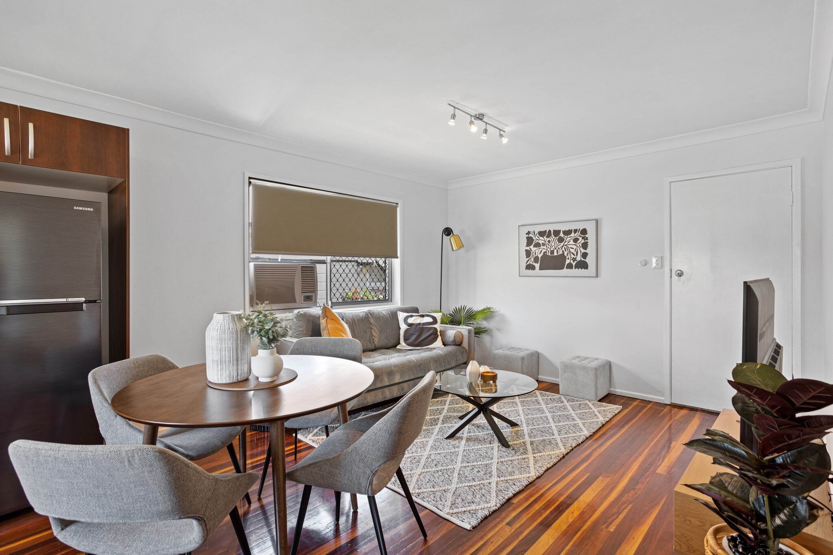 A modern living room with a dining area, a gray sofa, a coffee table, and a large window with blinds.