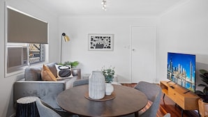 A modern living room with a round wooden table, grey sofas, a TV, and a cityscape painting. - Windsor Cosy 2-bedroom Unit in Quiet Neighborhood (Brisbane)
