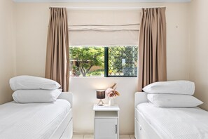 A bedroom with two single beds, a nightstand with a lamp and vase, and a window with curtains. - Albion Convenient Hideaway With Parking & Balcony (Brisbane)