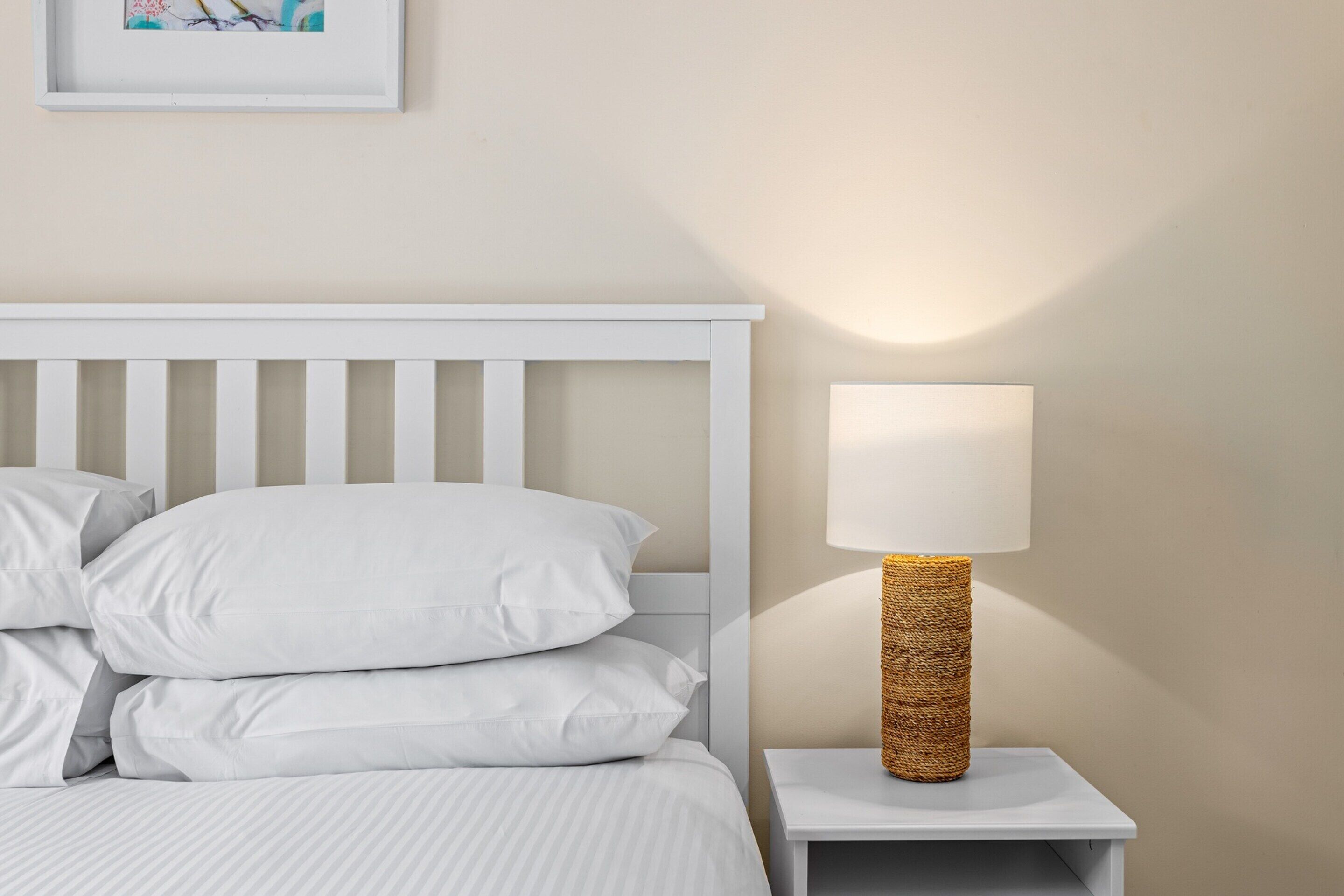 Una cama bien tendida con sábanas y almohadas blancas, una mesita de noche blanca con una lámpara de mimbre, y un cuadro en la pared.